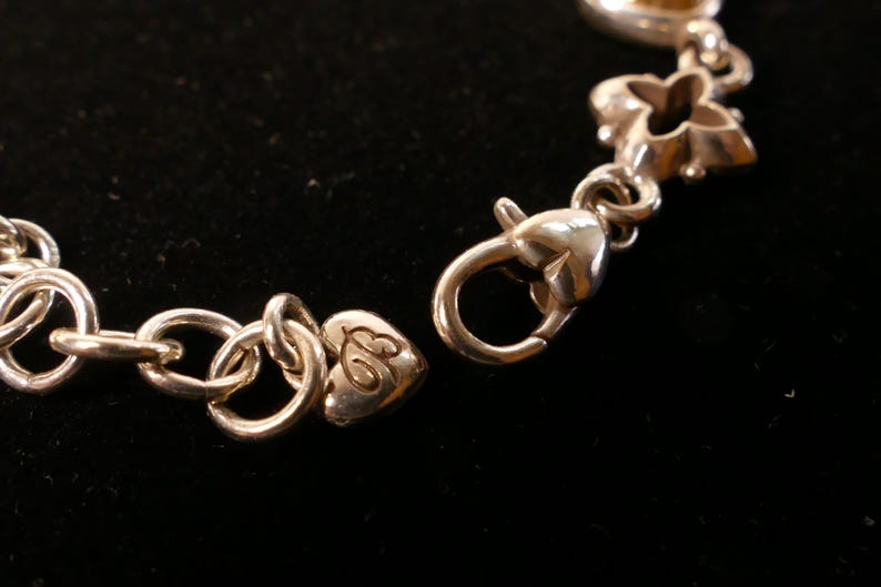 May include: Close-up of a silver bracelet with a heart-shaped clasp and decorative links. The bracelet is on a black background. The clasp is open, showing the connection point.