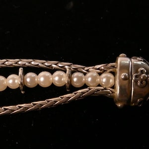 May include: Close-up of a silver necklace with a braided chain design. The necklace features a strand of white pearls and a decorative clasp. The background is black.
