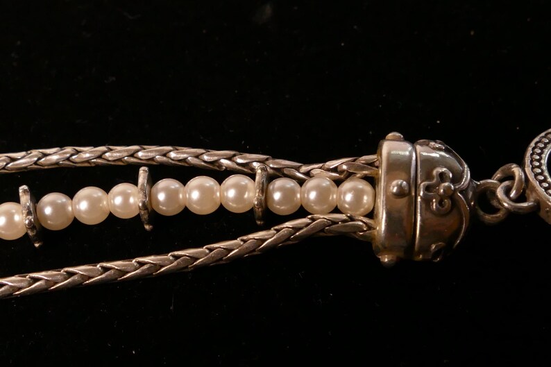 May include: Close-up of a silver necklace with a braided chain design. The necklace features a strand of white pearls and a decorative clasp. The background is black.