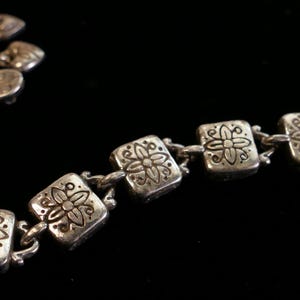 May include: A silver-toned bracelet with a floral design. The bracelet features square links with embossed flower patterns, connected by small chain links. The background is black, highlighting the metallic sheen of the jewelry.