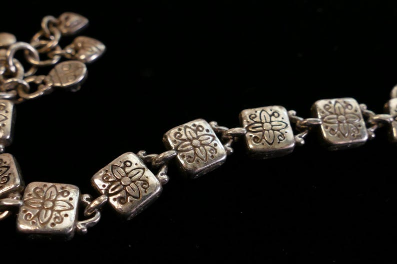 May include: A silver-toned bracelet with a floral design. The bracelet features square links with embossed flower patterns, connected by small chain links. The background is black, highlighting the metallic sheen of the jewelry.