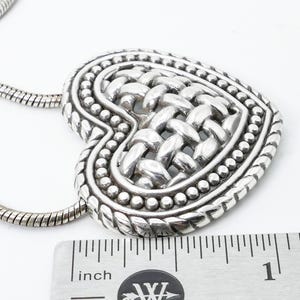 May include: A silver-toned heart pendant necklace. The heart features a woven design in the center, surrounded by a beaded border. The necklace chain is a snake chain style. The pendant measures approximately 1 inch in width.