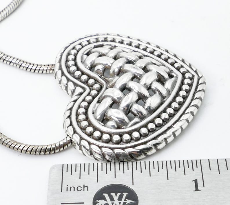 May include: A silver-toned heart pendant necklace. The heart features a woven design in the center, surrounded by a beaded border. The necklace chain is a snake chain style. The pendant measures approximately 1 inch in width.