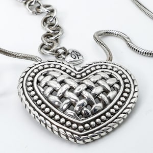 May include: Silver-toned heart pendant necklace. The pendant features a woven design in the center, surrounded by a beaded border and a braided edge. The necklace has a chain with a lobster clasp.