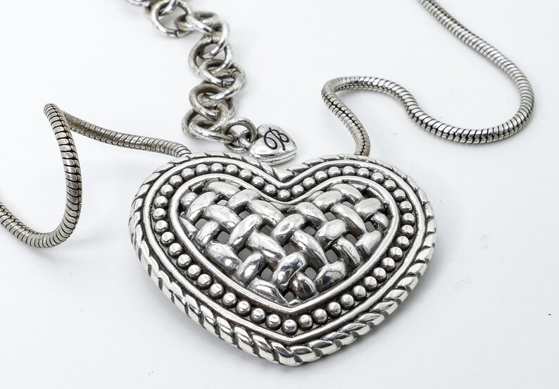 May include: Silver-toned heart pendant necklace. The pendant features a woven design in the center, surrounded by a beaded border and a braided edge. The necklace has a chain with a lobster clasp.