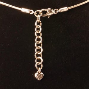 May include: Silver-toned necklace with a chain extender and lobster clasp. The extender chain has a small heart-shaped charm with a stylized letter "B" on it. The necklace is set against a black background.
