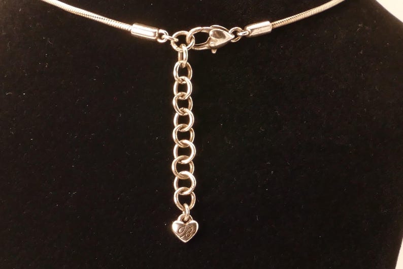 May include: Silver-toned necklace with a chain extender and lobster clasp. The extender chain has a small heart-shaped charm with a stylized letter "B" on it. The necklace is set against a black background.