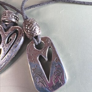 May include: Two silver-toned metal pendants on dark gray cord necklaces. One pendant is heart-shaped with cutouts, and the other is a rectangular tag with a heart cutout, engraved star motifs, and a textured silver bead above the tag.