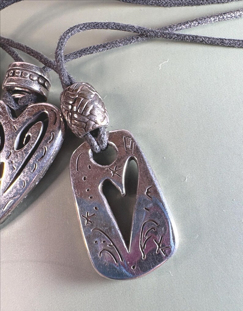 May include: Two silver-toned metal pendants on dark gray cord necklaces. One pendant is heart-shaped with cutouts, and the other is a rectangular tag with a heart cutout, engraved star motifs, and a textured silver bead above the tag.