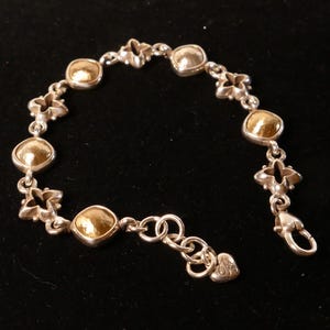 May include: Silver bracelet featuring gold-coloured, rounded square accents and floral-shaped links. The bracelet has a lobster clasp closure and a small heart-shaped charm. The jewellery is displayed on a black surface.