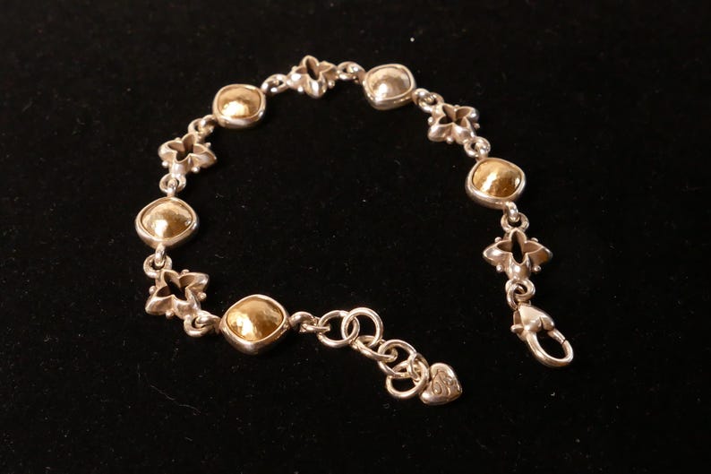 May include: Silver bracelet featuring gold-coloured, rounded square accents and floral-shaped links. The bracelet has a lobster clasp closure and a small heart-shaped charm. The jewellery is displayed on a black surface.