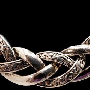 May include: Silver-toned necklace with a woven chain design. The links have a hammered texture and are adorned with intricate scrollwork. The necklace is set against a black background, highlighting its metallic sheen.
