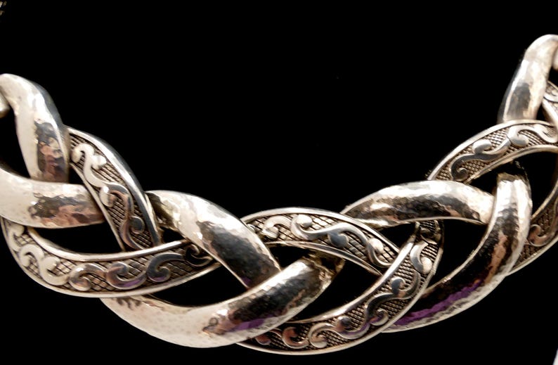 May include: Silver-toned necklace with a woven chain design. The links have a hammered texture and are adorned with intricate scrollwork. The necklace is set against a black background, highlighting its metallic sheen.