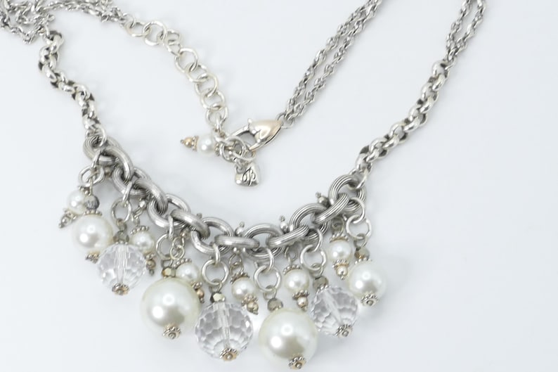 K&ouml;nnte beinhalten: Silberfarbene Halskette mit Kette und Anh&auml;nger mit Kunstperlen und klaren, facettierten Perlen. Die Halskette hat einen Karabinerverschluss und wird auf wei&szlig;em Hintergrund pr&auml;sentiert. Die Halskette ist ein Modeaccessoire.