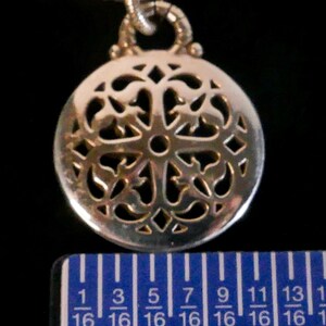 May include: A silver-toned circular pendant featuring an intricate openwork filigree pattern, suspended from a twisted metal chain. A blue ruler is positioned at the base, indicating the pendant has a diameter of approximately 0.75 inches.