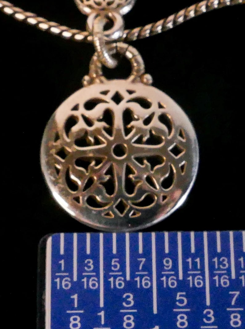 May include: A silver-toned circular pendant featuring an intricate openwork filigree pattern, suspended from a twisted metal chain. A blue ruler is positioned at the base, indicating the pendant has a diameter of approximately 0.75 inches.