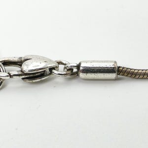 May include: Close-up of a silver-toned necklace clasp and chain. The clasp is heart-shaped, and the chain has a snake-like design. The necklace is on a white background.
