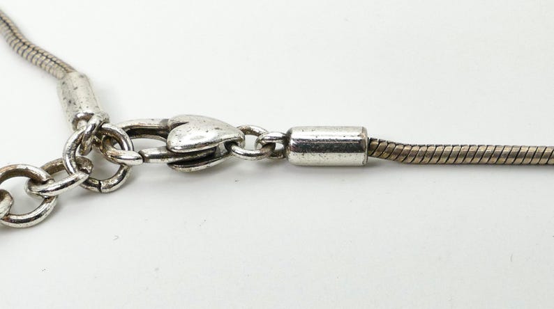 May include: Close-up of a silver-toned necklace clasp and chain. The clasp is heart-shaped, and the chain has a snake-like design. The necklace is on a white background.