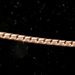 May include: A gold-toned metal chain necklace featuring a flat, interlocking snake-like link pattern. The chain has a smooth, reflective surface and a consistent width throughout its length, displaying a flexible and continuous design structure.