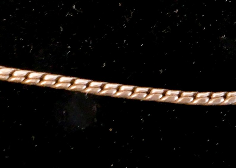 May include: A gold-toned metal chain necklace featuring a flat, interlocking snake-like link pattern. The chain has a smooth, reflective surface and a consistent width throughout its length, displaying a flexible and continuous design structure.