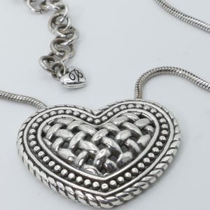 May include: Silver-toned heart pendant necklace. The heart-shaped pendant features a woven center design, surrounded by a beaded border and a rope-like edge. The necklace has a snake chain and an adjustable chain with a heart charm.