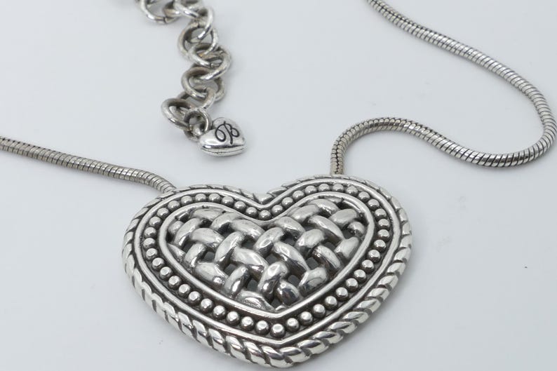 May include: Silver-toned heart pendant necklace. The heart-shaped pendant features a woven center design, surrounded by a beaded border and a rope-like edge. The necklace has a snake chain and an adjustable chain with a heart charm.