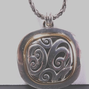 May include: Silver-tone pendant necklace with a decorative swirl design. The pendant has a gold-tone inner frame and a braided chain. The pendant is a square shape with rounded edges.