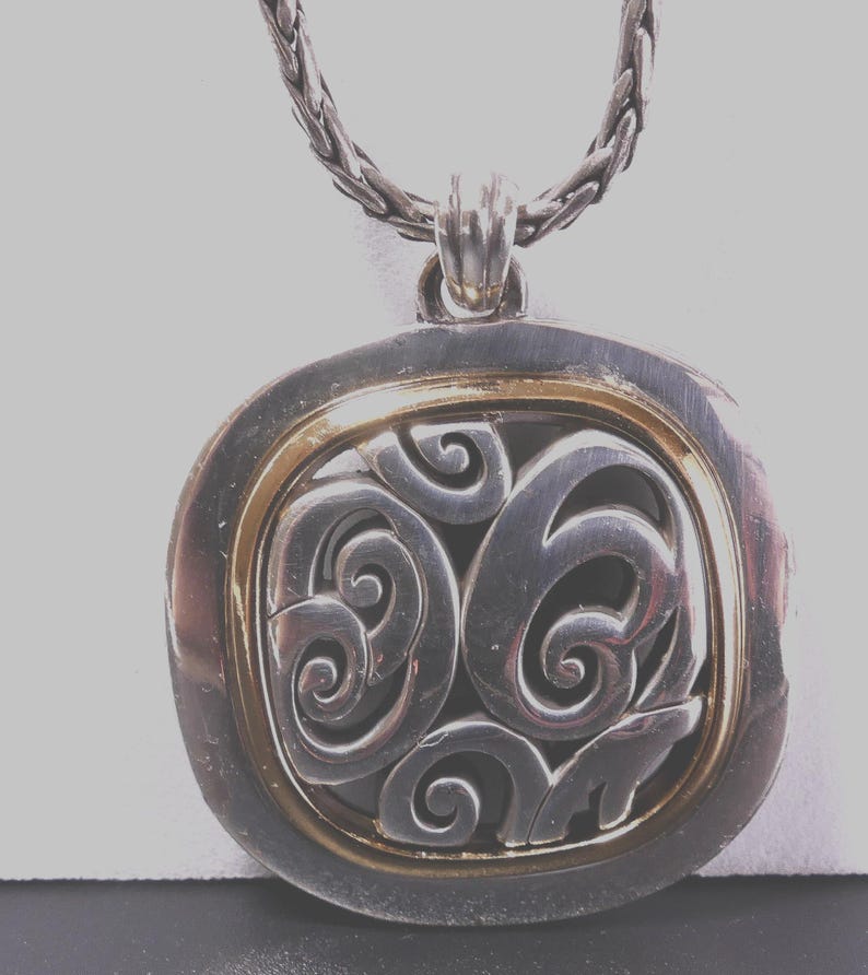 May include: Silver-tone pendant necklace with a decorative swirl design. The pendant has a gold-tone inner frame and a braided chain. The pendant is a square shape with rounded edges.