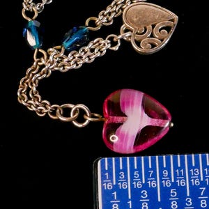 Puede incluir: Un collar de cadena plateada con un colgante de coraz&oacute;n de vidrio rosa y blanco, un dije de coraz&oacute;n de plata con filigrana recortada y cuentas facetadas de color azul oscuro. Una regla azul muestra las medidas en cent&iacute;metros.