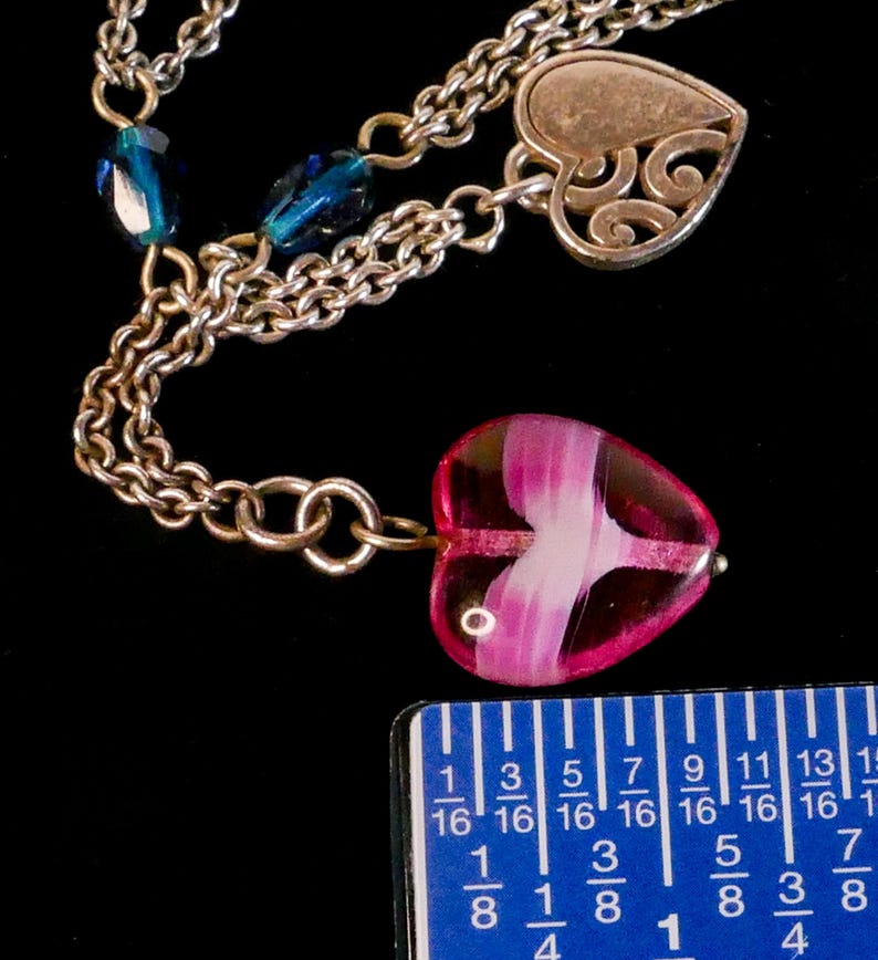 Puede incluir: Un collar de cadena plateada con un colgante de coraz&oacute;n de vidrio rosa y blanco, un dije de coraz&oacute;n de plata con filigrana recortada y cuentas facetadas de color azul oscuro. Una regla azul muestra las medidas en cent&iacute;metros.