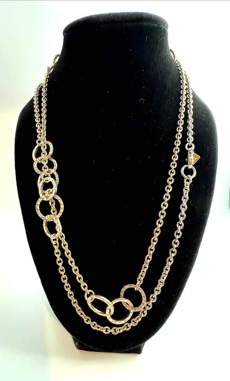 Puede incluir: Collar dorado con dos hilos y una serie de c&iacute;rculos entrelazados. El collar se exhibe en un maniqu&iacute; negro. Los c&iacute;rculos tienen un acabado texturizado y la cadena es un dise&ntilde;o de eslabones delicados.