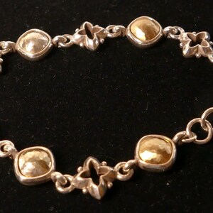 May include: Silver bracelet with gold-toned square and silver quatrefoil links. The bracelet has a lobster clasp. The jewellery is displayed on a black surface. The bracelet's design is symmetrical.