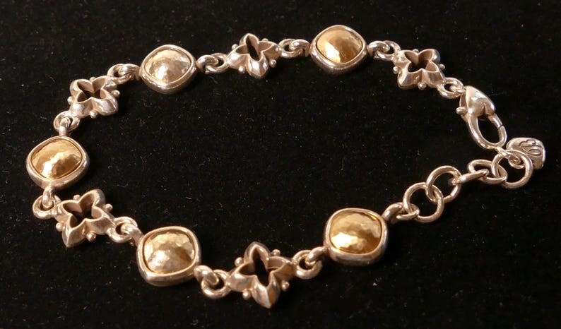 May include: Silver bracelet with gold-toned square and silver quatrefoil links. The bracelet has a lobster clasp. The jewellery is displayed on a black surface. The bracelet's design is symmetrical.