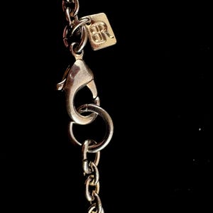 Puede incluir: Primer plano de un cierre y cadena de collar plateados sobre un fondo negro. Una peque&ntilde;a etiqueta cuadrada con las letras "BR" est&aacute; unida a la cadena. Los eslabones de la cadena son peque&ntilde;os y delicados.