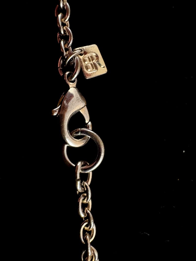 Puede incluir: Primer plano de un cierre y cadena de collar plateados sobre un fondo negro. Una peque&ntilde;a etiqueta cuadrada con las letras "BR" est&aacute; unida a la cadena. Los eslabones de la cadena son peque&ntilde;os y delicados.