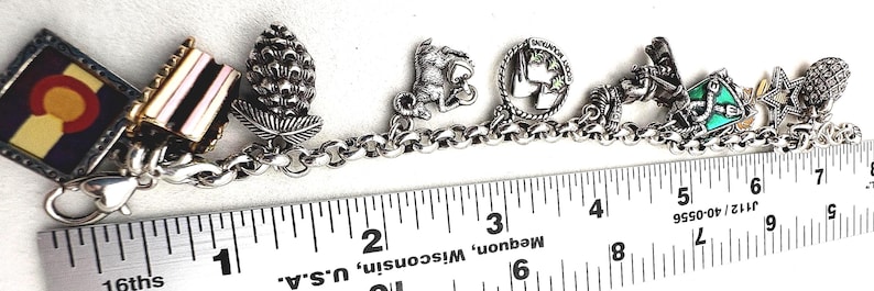 May include: A silver charm bracelet with various detailed charms, including a Colorado flag, a pinecone, and a squirrel. The bracelet is measured with a ruler, showing a length of approximately 7 inches.