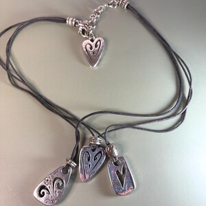 May include: A multi-strand dark gray cord necklace featuring three silver-toned metal pendants with cut-out heart and floral designs, plus a small heart-shaped charm near the silver-toned metal lobster clasp and adjustable chain extender.