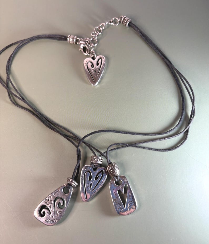 May include: A multi-strand dark gray cord necklace featuring three silver-toned metal pendants with cut-out heart and floral designs, plus a small heart-shaped charm near the silver-toned metal lobster clasp and adjustable chain extender.