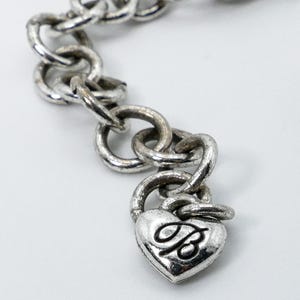 May include: A silver-toned chain bracelet with a heart-shaped charm. The charm features a stylized letter "B" and a padlock design. The bracelet links are circular and connected. The background is white.