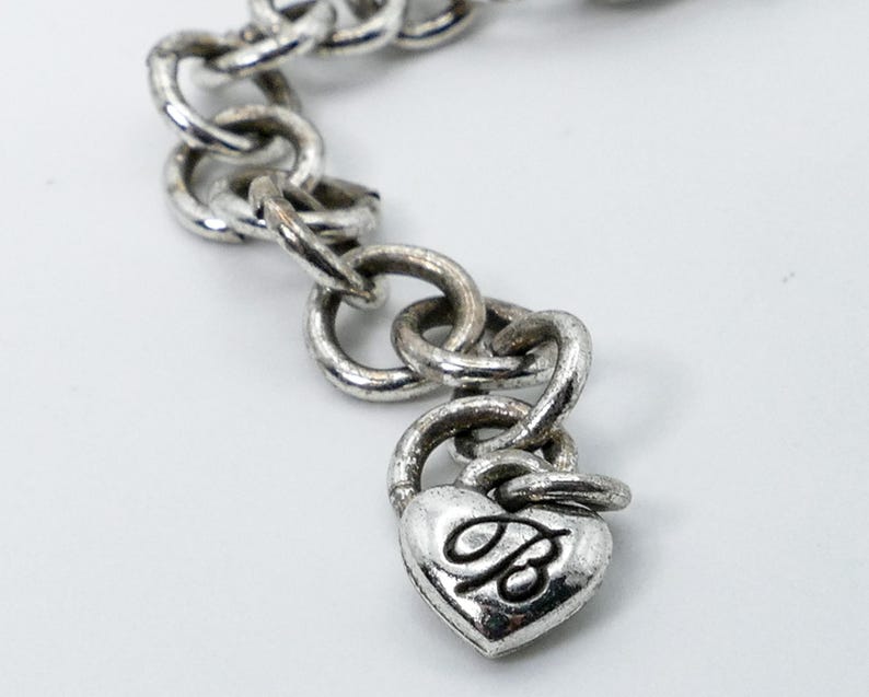 May include: A silver-toned chain bracelet with a heart-shaped charm. The charm features a stylized letter "B" and a padlock design. The bracelet links are circular and connected. The background is white.