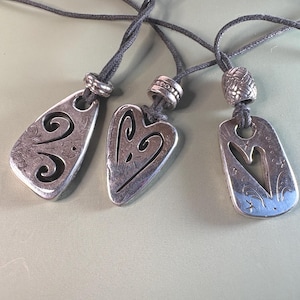 May include: Three silver-toned metal pendants on dark gray cords. Each pendant features a unique cutout design, including two heart-shaped motifs and one abstract swirl pattern. Each cord is accented with a textured silver-toned metal bead.