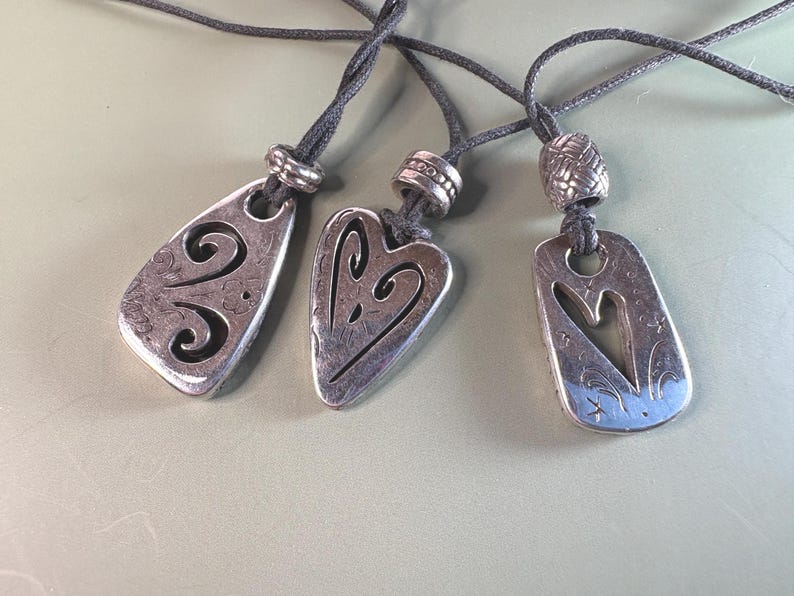May include: Three silver-toned metal pendants on dark gray cords. Each pendant features a unique cutout design, including two heart-shaped motifs and one abstract swirl pattern. Each cord is accented with a textured silver-toned metal bead.