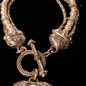 May include: Silver-toned bracelet featuring a toggle clasp and a heart-shaped pendant. The bracelet has multiple strands of small, round pearls and a decorative heart pendant with a single pearl in the center. The bracelet is on a black background.