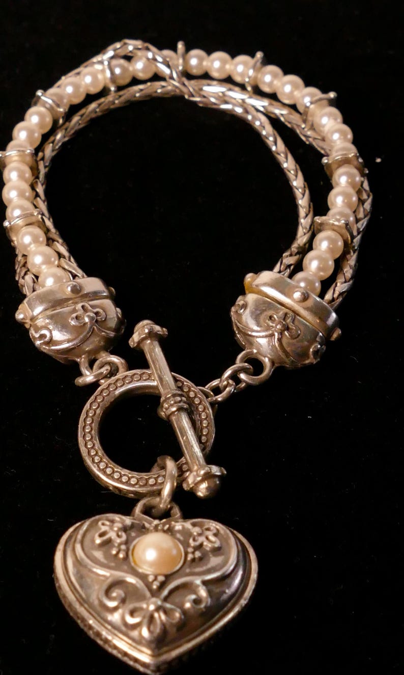 May include: Silver-toned bracelet featuring a toggle clasp and a heart-shaped pendant. The bracelet has multiple strands of small, round pearls and a decorative heart pendant with a single pearl in the center. The bracelet is on a black background.