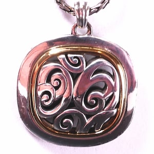 May include: Silver-tone pendant necklace with a gold-tone inner frame. The pendant features an openwork design of swirling shapes. The necklace chain is a braided style. The pendant is square with rounded corners.
