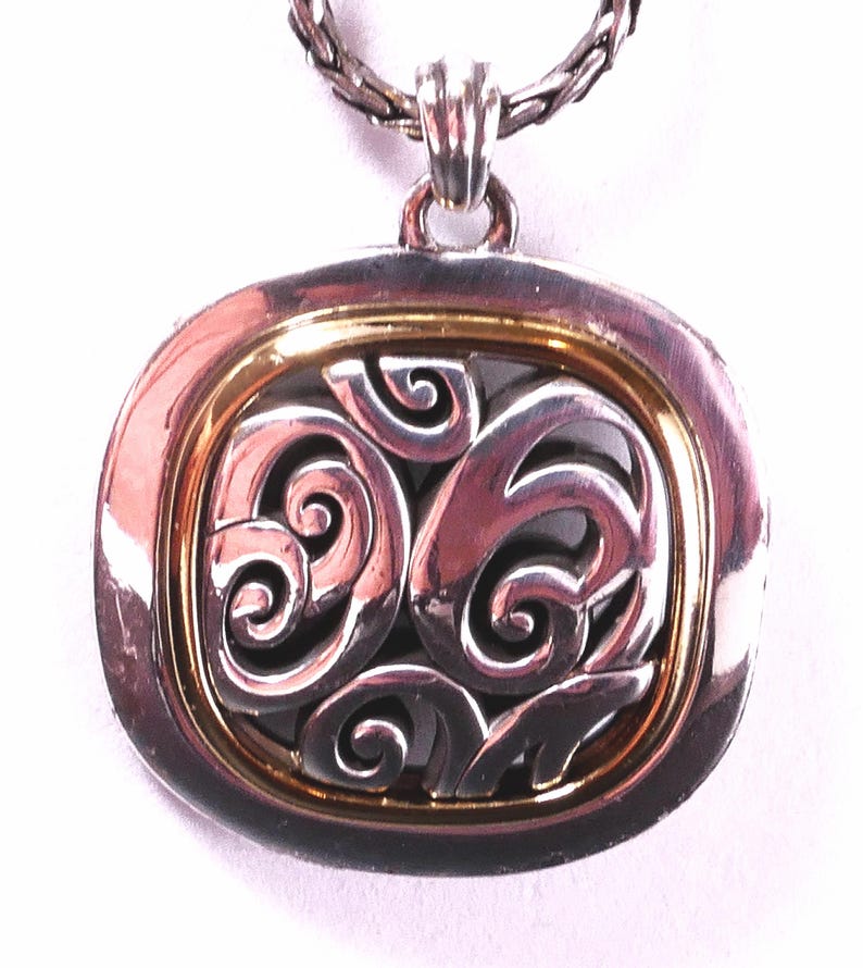 May include: Silver-tone pendant necklace with a gold-tone inner frame. The pendant features an openwork design of swirling shapes. The necklace chain is a braided style. The pendant is square with rounded corners.