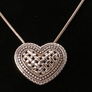 May include: Silver heart pendant necklace. The heart-shaped pendant features a woven center design, surrounded by a beaded border. The necklace has a delicate chain. The jewelry is displayed against a black background.