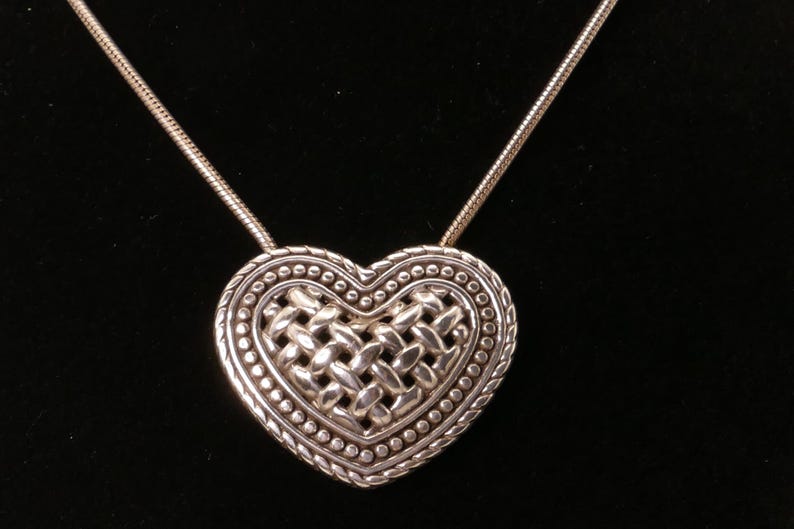 May include: Silver heart pendant necklace. The heart-shaped pendant features a woven center design, surrounded by a beaded border. The necklace has a delicate chain. The jewelry is displayed against a black background.