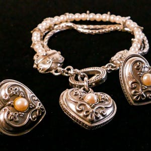 May include: A silver bracelet with pearl accents and heart-shaped charms, each featuring a central pearl. The set includes matching heart-shaped earrings. The jewelry is displayed against a black background.