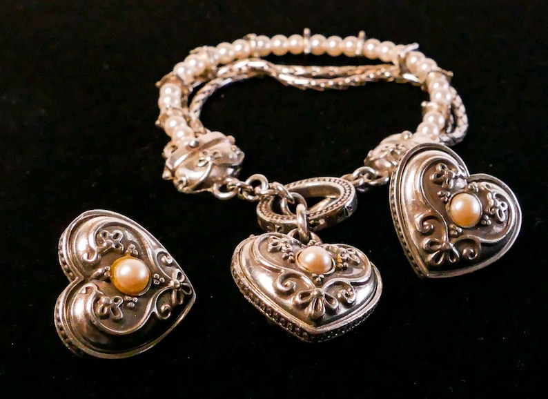 May include: A silver bracelet with pearl accents and heart-shaped charms, each featuring a central pearl. The set includes matching heart-shaped earrings. The jewelry is displayed against a black background.